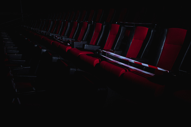 red chairs in the cinema hall are illuminated by a beam of light, a low key, a restrictive tape, quarantine. Selective focus. Empty cinema, quarantine COVID-19, SARS-CoV-2, 2019-nCoV - Photo, Image