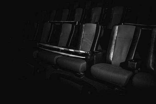 červené židle v kinosálu jsou osvětleny paprskem světla, nízkým klíčem, omezující páskou, karanténou. Selektivní soustředění. Prázdné kino, karanténa COVID-19, SARS-CoV-2, 2019-nCoV - Fotografie, Obrázek