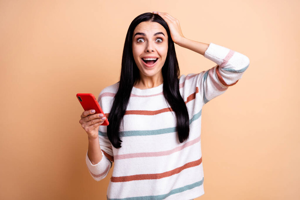 Photo of young crazy woman happy smile amazed shocked surprised use cellphone hand touch head isolated over beige color background - Fotoğraf, Görsel