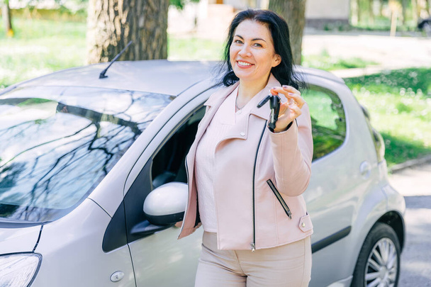 幸せな女性ドライバーは車のキーを示し、オープンカーのドアの近くに立っています。彼女は購入に満足しています - 写真・画像