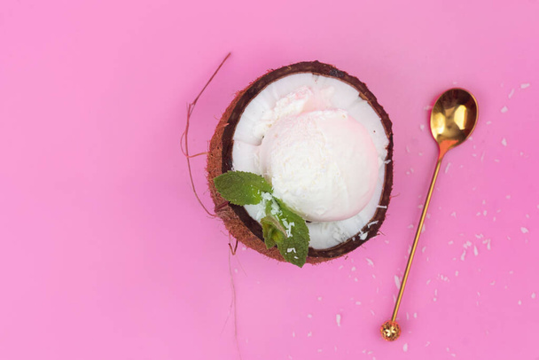 Vanilla ice cream balls in a fresh coconut half decorated with mint leaves with a spoon on a pink background. - Фото, изображение