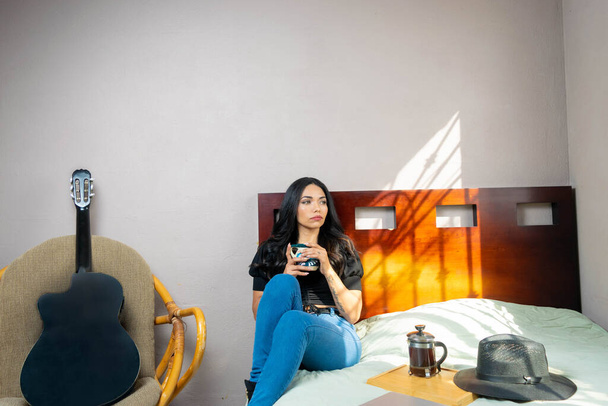 bela mulher latina descansando sentado na cama bebendo café mexicano em copo colorido ao lado da imprensa francesa e chapéu - Foto, Imagem