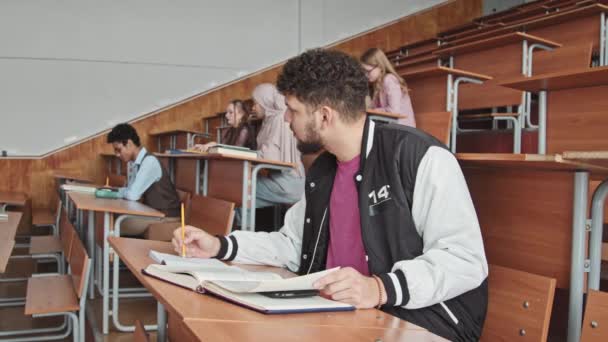 Side-view medium slowmo of young male student sitting by desk in auditorium trying to cheat at at exam, looking at smartphone put between pages of student book - Záběry, video