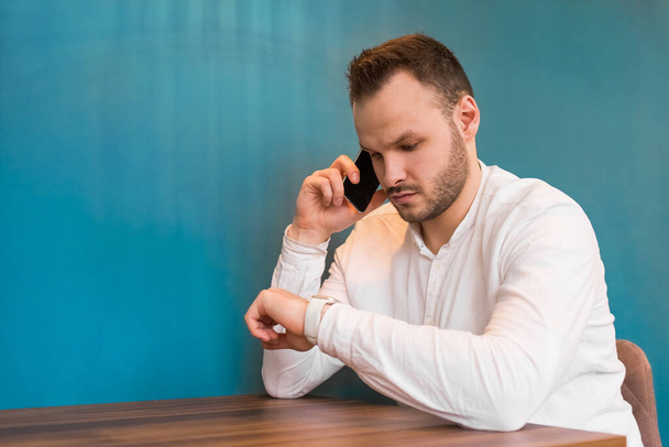 A young European guy businessman in a white shirt, with a beard sits at a table, talks on a mobile phone and looks at the time on a blue background. - 写真・画像