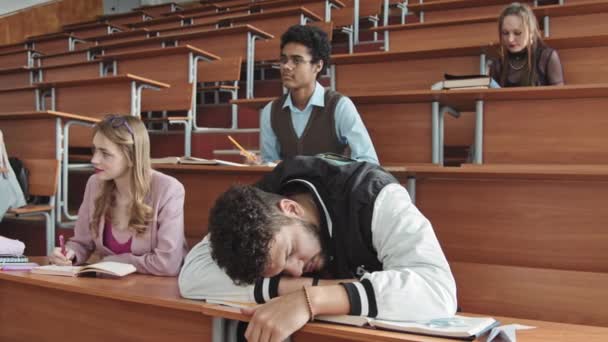Medium shot of lazy male student in sports bomber jacket sleeping on desk in lecture hall or auditorium while his Mixed-race classmate poking at him with pencil to wake up as professor entering - Footage, Video