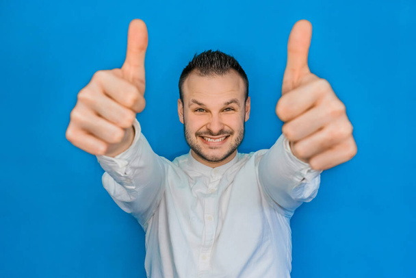 Portré fiatal vonzó európai boldog üzletember fehér ingben szakállas mutatja hűvös hüvelykujj fel két kézzel a kék háttér. - Fotó, kép