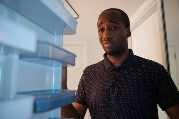 An African American boy opens the fridge and realizes it is empty. He has a sad and worried facial expression. Crisis and poverty concept. - Фото, изображение