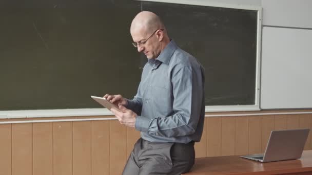 Retrato lento médio de professor universitário adulto médio em negrito em trajes casuais inteligentes e óculos posando para câmera com tablet digital em mãos em pé no quadro negro - Filmagem, Vídeo