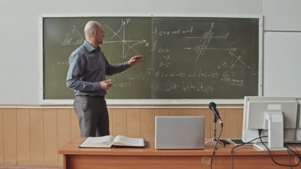 Średnie spowolnienie pewnego siebie męskiego profesora uniwersytetu średniej wielkości stojącego przy tablicy w audytorium lub sali wykładowej, wyjaśniające studentom, jak rozwiązać problemy z geometrią twardą - Materiał filmowy, wideo