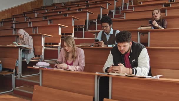 Lentitud media de un pequeño grupo de jóvenes universitarios multiétnicos o estudiantes de secundaria sentados junto a largos escritorios de madera en filas y desplazándose a través de sus teléfonos inteligentes durante la lección - Metraje, vídeo