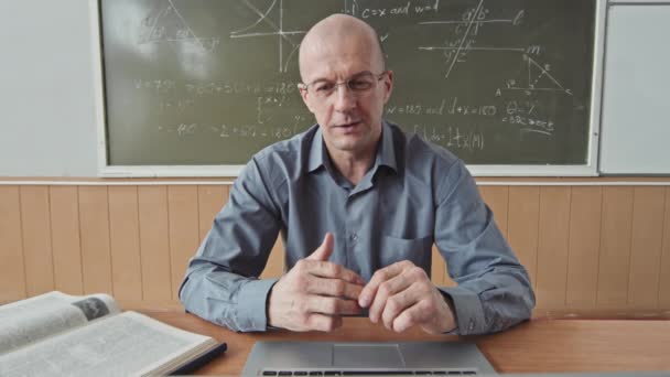 POV plan moyen de professeur masculin confiant mi-adulte enseignant les mathématiques parlant à la caméra assis sur le bureau au tableau noir dans une salle de classe lumineuse - Séquence, vidéo