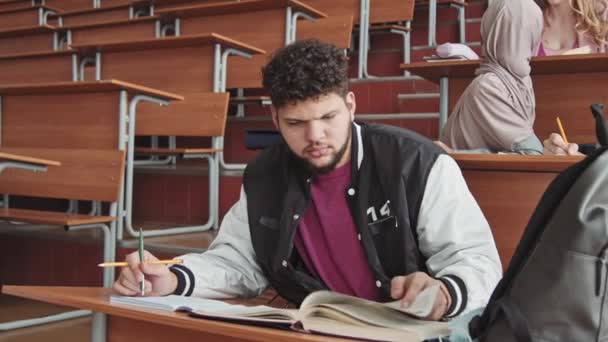Média lentidão de jovem estudante barbudo em esportes bomber jaqueta virando páginas de livro de estudante grosso sentado à mesa em grande auditório com seus colegas multiétnicos - Filmagem, Vídeo