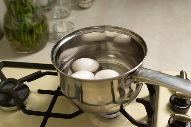 Белые куриные яйца варят в ковше с ватой на плите в домашней кухне. Концепция диетического питания. Горизонтальная ориентация. - Фото, изображение