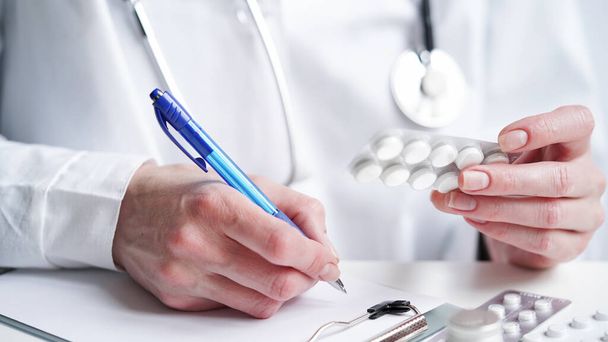 Close-up of a doctor writing a prescription in a medical record on a tablet. A doctor in a white coat holds pills in his hands and writes a prescription with a blue pen. Medical banner concept - Foto, imagen