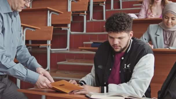 Mittlere Verlangsamung eines jungen bärtigen lateinamerikanischen Studenten in Bomberjacke, der sich Notizen in einem Textbuch von einem erwachsenen Professor ansieht, der ihm bei Test oder Prüfung hilft. - Filmmaterial, Video