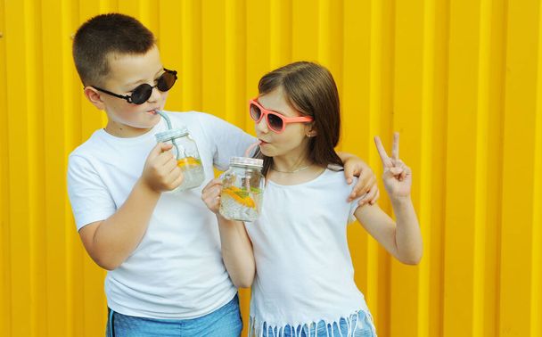 Little friends in white t-shirts hold summer cold cocktails with orange and mint on a yellow background - Φωτογραφία, εικόνα