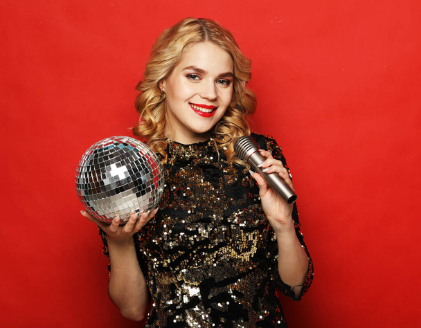 Christmas, celebration party, holiday concept. Young woman in evening dress holding microphone and disco ball. Brighrt make up and Wavy hairstyle. Party time. - Zdjęcie, obraz