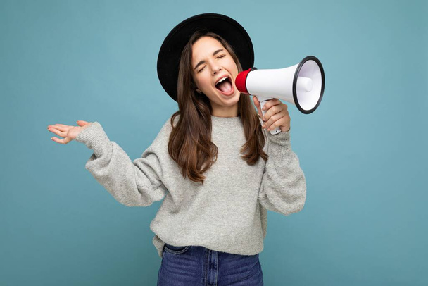 Photo of young beautiful positive emotional brunette woman with sincere emotions wearing stylish black hat and gray sweater isolated over blue background with copy space and shouting into magaphone - Foto, afbeelding