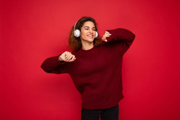 Photo of beautiful happy smiling young brunette curly woman wearing dark red sweater isolated over red background wall wearing white bluetooth headphones listening to music and having fun looking to - Foto, Imagen