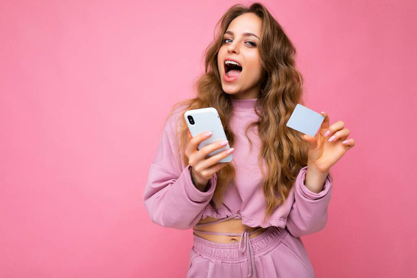 Photo of attractive overjoyed adult blonde curly woman wearing pink clothes isolated over pink background using mobile phone making payment using credit card looking at camera and having fun - Foto, imagen
