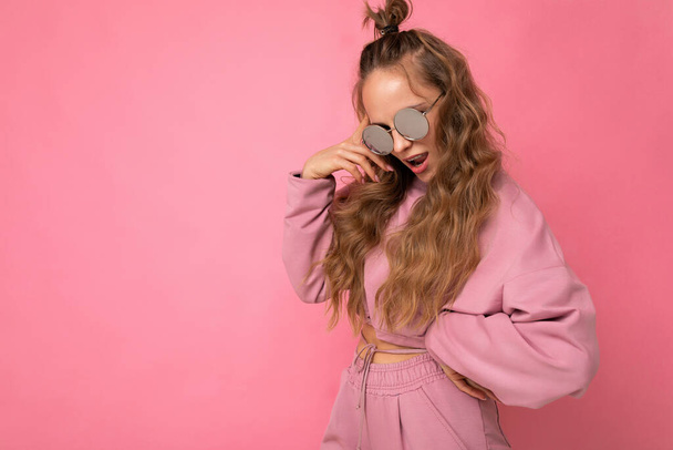Shot of beautiful happy young dark blonde curly woman isolated over pink background wall wearing casual pink sport clothes and stylish sunglasses looking at camera - Zdjęcie, obraz