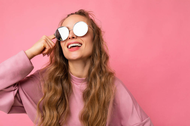 Shot of beautiful happy smiling young dark blonde curly woman isolated over pink background wall wearing casual pink sport clothes and stylish sunglasses looking to the side - Fotoğraf, Görsel