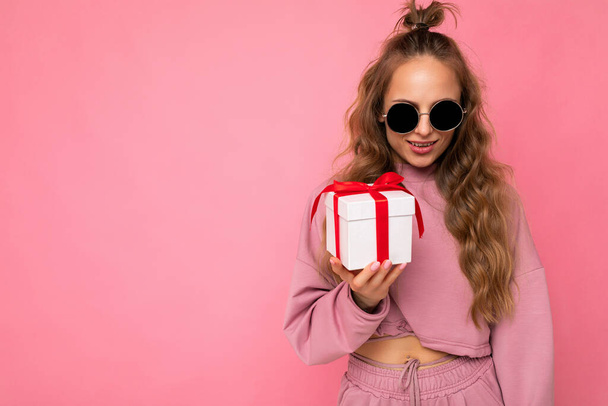 Приваблива позитивна молода блондинка кучерява жінка ізольована на рожевій стіні в рожевому спортивному одязі та сонцезахисних окулярах, що тримає подарункову коробку, дивлячись на камеру
 - Фото, зображення