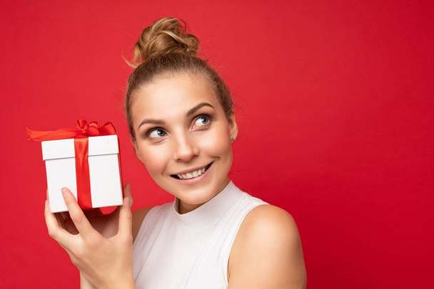 Beautiful happy young blonde woman isolated over colourful background wall wearing stylish casual clothes holding gift box and looking to the side - Photo, Image