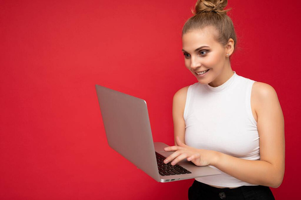 Seitenprofil-Foto von schönen überglücklichen blonden Frau mit gerafften Haaren trägt weißes T-Shirt mit Computer-Laptop-Eingabe auf der Tastatur Blick auf Netbook-Monitor isoliert über rot - Foto, Bild