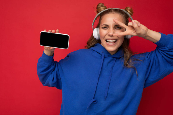 Aantrekkelijke positieve glimlachende jonge vrouw in stijlvolle casual outfit geïsoleerd op kleurrijke achtergrond wandhouder en toont mobiele telefoon met leeg scherm voor cutout dragen van witte bluetooth - Foto, afbeelding