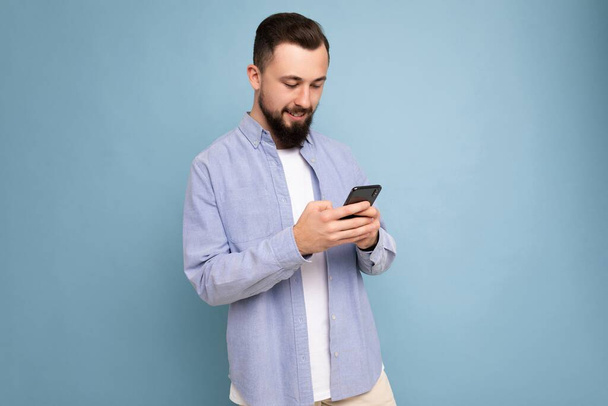 Photo shot of handsome positive good looking young man wearing casual stylish outfit poising isolated on background with empty space holding in hand and using mobile phone messaging sms looking at - Photo, Image