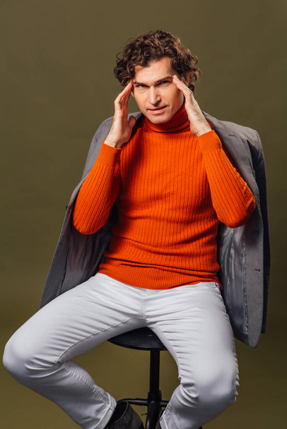 Portrait of a tall handsome man dressed in orange turtleneck, grey jacket and white jeans posing on the green background showing headache - 写真・画像