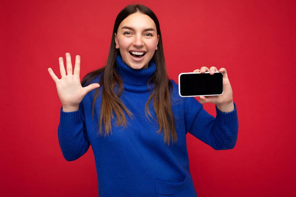 Detailní portrét fotografie záběr okouzlující mladé brunetky žena na sobě ležérní modrý svetr izolované přes červené pozadí s prázdným prostorem v ruce mobilní telefon a ukazující smartphone s - Fotografie, Obrázek