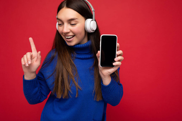 Atrakcyjny szczęśliwy uśmiechnięty młoda brunetka kobieta w niebieskim swetrze odizolowany na czerwonym tle gospodarstwa i korzystania z telefonu komórkowego surfing w Internecie w białe słuchawki słuchanie muzyki patrząc - Zdjęcie, obraz