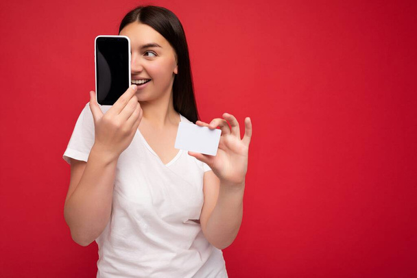 Szekrény fotó Elég boldog fiatal barna nő visel alkalmi fehér póló elszigetelt piros háttér üres hely tartja a kezében mobiltelefon és mutatja okostelefon üres képernyőn - Fotó, kép