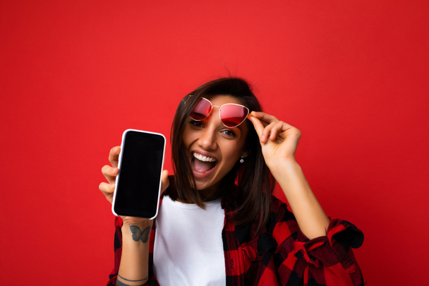 Fénykép gyönyörű mosolygós fiatal nő csinos visel alkalmi elegáns ruhát áll elszigetelt a háttérben másolás tér gazdaság okostelefon mutatja, telefon a kezében üres képernyő kijelző - Fotó, kép