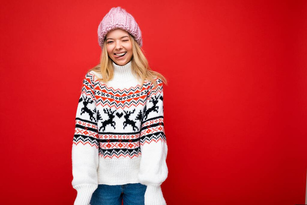 Foto di una giovane donna bionda piuttosto positiva isolata sul muro di sfondo rosso che indossa maglione invernale e cappello rosa guardando la fotocamera - Foto, immagini