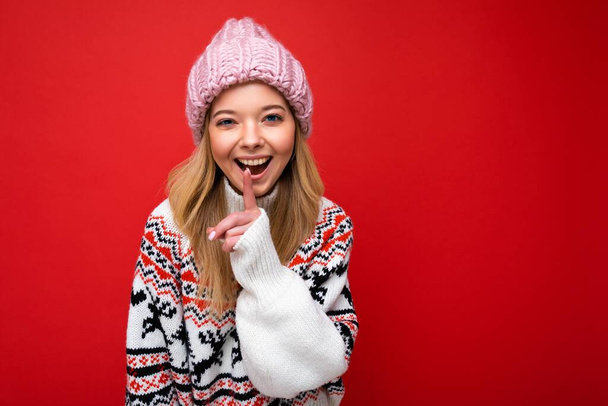 Портрет молодої позитивної щасливої привабливої блондинки з щирими емоціями в рожевому капелюсі і зимовий светр ізольовані на червоному тлі з копіювальним простором і показуючи жест
 - Фото, зображення