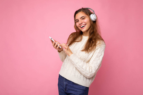 Φωτογραφία από την όμορφη χαρούμενη χαμογελαστή νεαρή γυναίκα φορώντας κομψό casual στολή που απομονώνονται πάνω από πολύχρωμο φόντο τοίχο φορώντας λευκό bluetooth ασύρματα ακουστικά και ακούγοντας μουσική και - Φωτογραφία, εικόνα