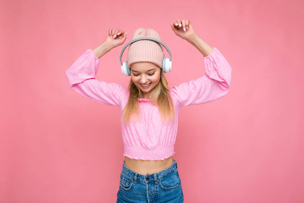 Gyönyörű boldog mosolygós fiatal szőke nő visel rózsaszín blúz és rózsaszín kalap elszigetelt felett rózsaszín háttér fal visel fehér vezeték nélküli bluetooth fejhallgató hallgat hűvös zene szórakozás és - Fotó, kép