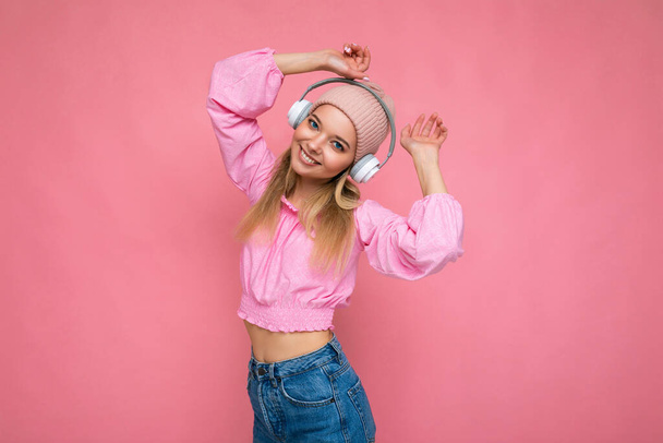 Foto di attraente positivo sorridente giovane donna bionda indossa camicetta rosa e cappello rosa isolato sopra parete sfondo rosa indossando auricolari bluetooth wireless bianchi ascoltare musica divertirsi e - Foto, immagini