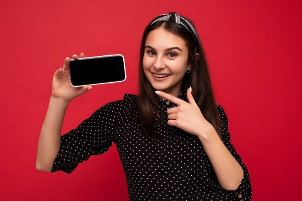 Closeup photo of happy Pretty brunette girl standing isolated over red wall wearing casual stylish black clothes showing mobile phone with empty screen for cutout looking at camera and pointing finger - Photo, Image