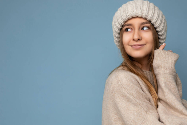 Shot of beautiful sexy smiling adult blonde woman standing isolated on blue background wall wearing beige warm sweater and winter beige hat looking to the side and enjoying - Photo, image