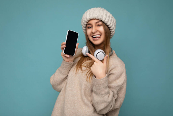 Photo sexy attrayant positif joyeux jeune femme blonde sombre portant un pull élégant beige et tricoté chapeau d'hiver beige isolé sur fond bleu portant des écouteurs sans fil bluetooth blanc et - Photo, image