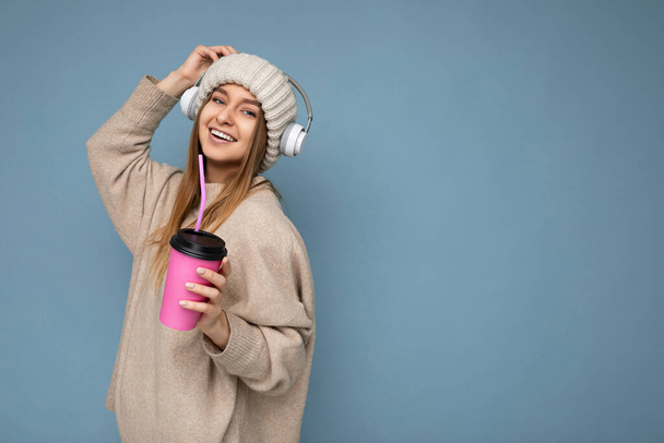 Side-profile photo of Beautiful happy lachende jonge blonde vrouw draagt beige winter trui en hoed geïsoleerd over blauwe achtergrond drinken drank dragen witte draadloze bluetooth headsets - Foto, afbeelding