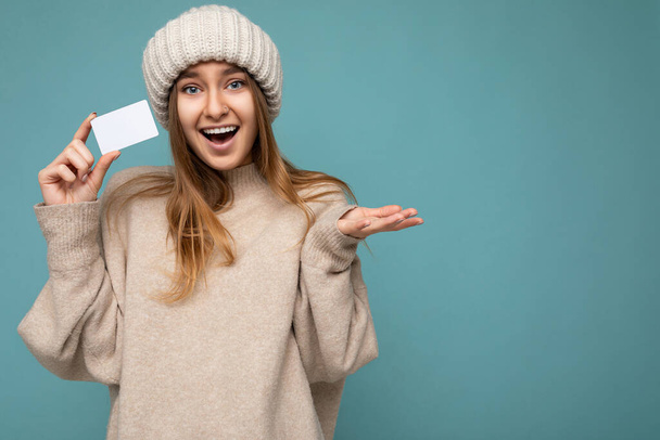 Photo shot of Beautiful amazed positive smiling young dark blonde woman wearing beige sweater and knitted beige hat isolated over blue background holding and showing bank credit card looking at camera - Photo, Image