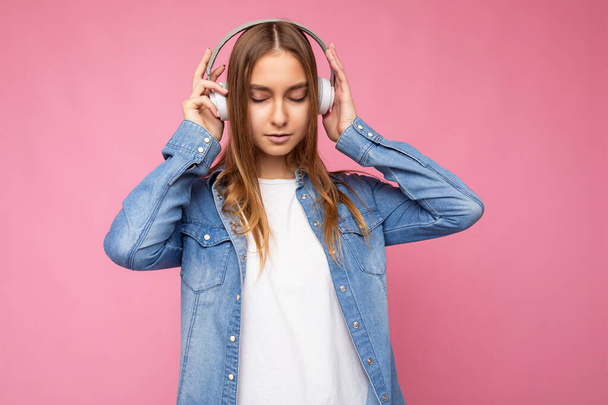 Schön mit aufrichtigen Emotionen junge blonde Frau trägt blaues Jeanshemd und weißes T-Shirt isoliert über rosa Hintergrund tragen weiße drahtlose Bluetooth-Kopfhörer hören coole Musik und - Foto, Bild