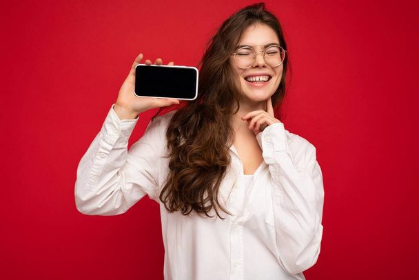 Smiling attractive young brunet woman wearing white shirt and optical glasses isolated over red background holding in hand and showing mobile phone with empty screen for mockup and having fun - Foto, immagini