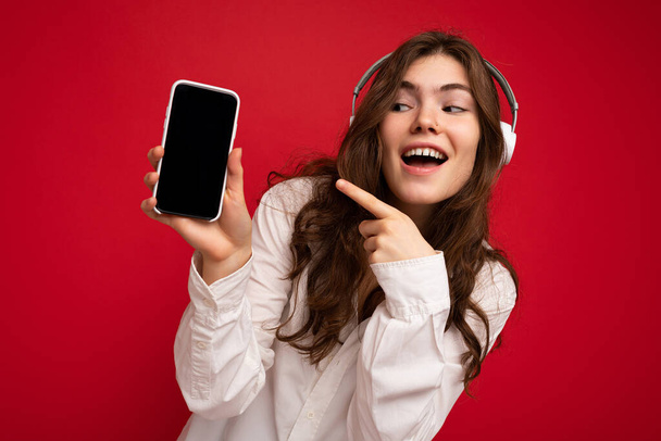 Atrakcyjne sexy sexy młody brunetka kręcone kobieta noszenie białą koszulę izolowane na czerwonym tle ściany gospodarstwa i pokazuje telefon komórkowy z pustym wyświetlaczem w białym bluetooth słuchawki słuchanie - Zdjęcie, obraz