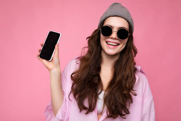 Kuva kaunis hymyilevä nuori nainen hyvännäköinen yllään rento tyylikäs asu seisoo eristetty taustalla kopio tilaa tilalla älypuhelin osoittaa puhelimen kädessä tyhjä näyttö - Valokuva, kuva